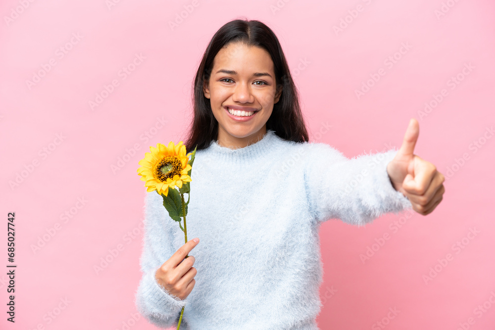 Young Colombian woman holding sunflower isolated on pink background giving a thumbs up gesture Young Colombian woman holding sunflower isolated on pink background giving a thumbs up gesture