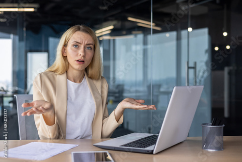 Free Download Portrait of an upset and disappointed businesswoman sitting in the office at the table, working on a laptop, looking seriously at the camera and spreading her hands, thinking about solving a problem - GFXLoad