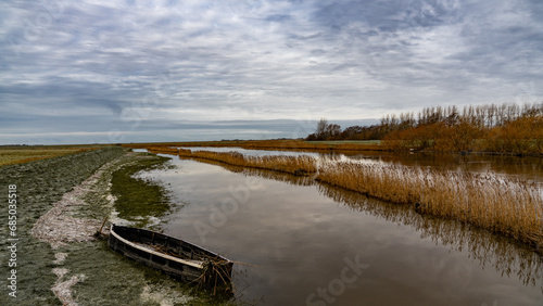 Low clouds hanging over a small river with an old boat on its bank that runs through fields in the Wadden Sea National Park