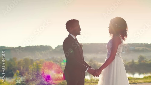 African American couple holding hands and talking romantic sentimental vows to each other in front of sunny landscape. Happy people in wedding outfit celebrating holiday of their love at nature. photo