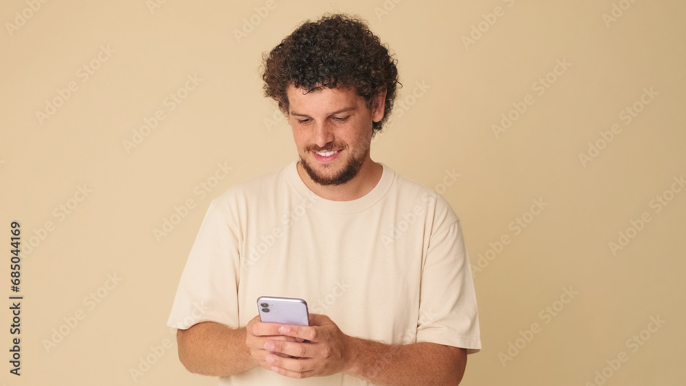 Laughing guy with curly hair dressed in beige t-shirt, uses mobile phone, browses social networks, funny videos standing in studio on beige background in the studio