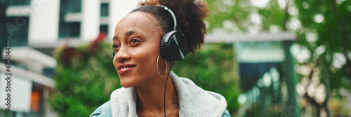 Close-up cute African girl with ponytail wearing denim jacket, in crop top with national pattern, listening to music with headphones in modern building background. photo