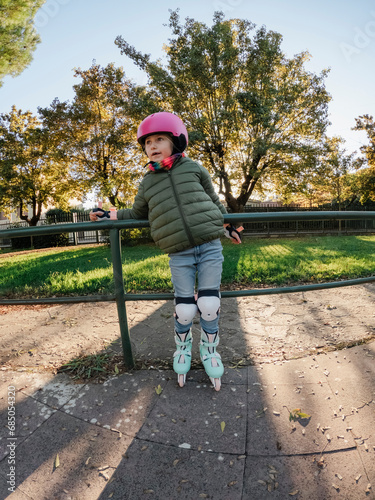 A child in a pink helmet and green jacket is rollerblading in a park, with trees and a sunset in the background.