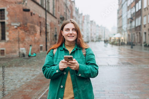 Attractive young female tourist with backpack is exploring new city. Redhead girl looking at camera and holding a smart phone on urban background. Traveling Europe in autumn. Vacation concept © mdyn