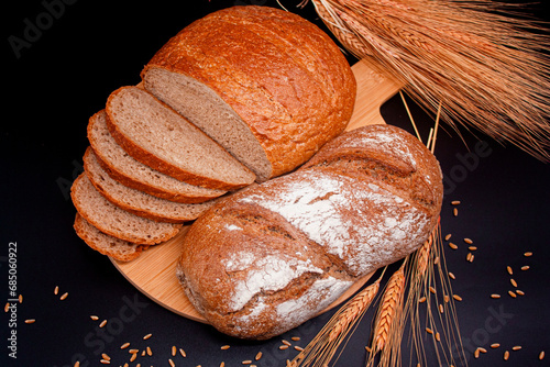 Whole and sliced breads, wheat ears and scattered wheat grains on wooden plate on black background. Photograph. (ID: 685060922)