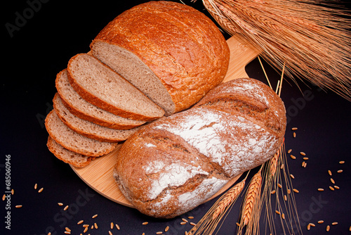 Whole and sliced breads, wheat ears and scattered wheat grains on wooden plate on black background. Photograph. (ID: 685060946)