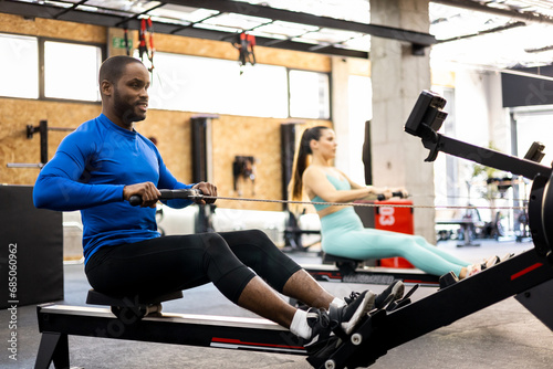 A couple of athletes of different ethnicities are working their bodies with a magnetic air rowing machine. The adult is in the foreground performing a back exercise with the magnetic air row.