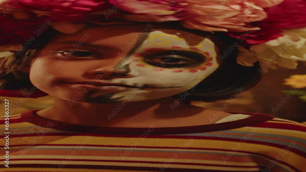 Vertical medium closeup portrait of little Hispanic girl with half painted face and flower wreath on her head posing with craft skeleton mask in backyard at night during Day of the Dead celebration