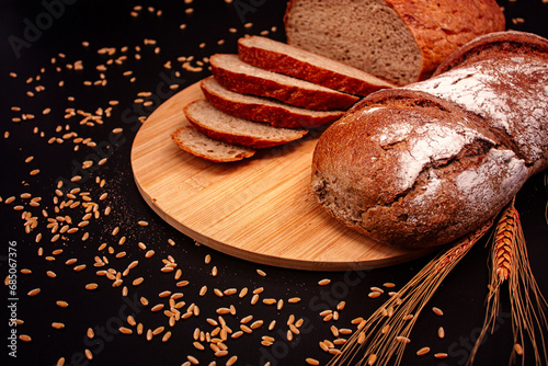 Whole and sliced breads, wheat ears and scattered wheat grains on wooden plate on black background. Photograph. (ID: 685067376)