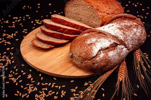 Whole and sliced breads, wheat ears and scattered wheat grains on wooden plate on black background. Photograph. (ID: 685067398)