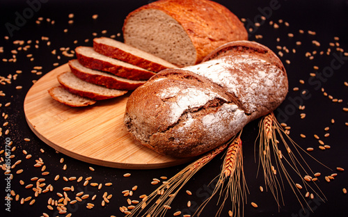 Whole and sliced breads, wheat ears and scattered wheat grains on wooden plate on black background. Photograph. (ID: 685067700)