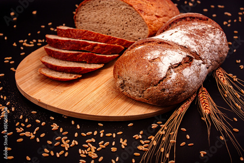 Whole and sliced breads, wheat ears and scattered wheat grains on wooden plate on black background. Photograph. (ID: 685068558)
