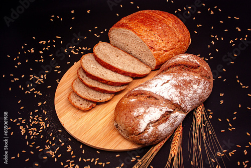 Whole and sliced breads, wheat ears and scattered wheat grains on wooden plate on black background. Photograph. (ID: 685069188)