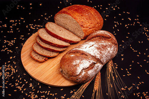 Whole and sliced breads, wheat ears and scattered wheat grains on wooden plate on black background. Photograph. (ID: 685069363)
