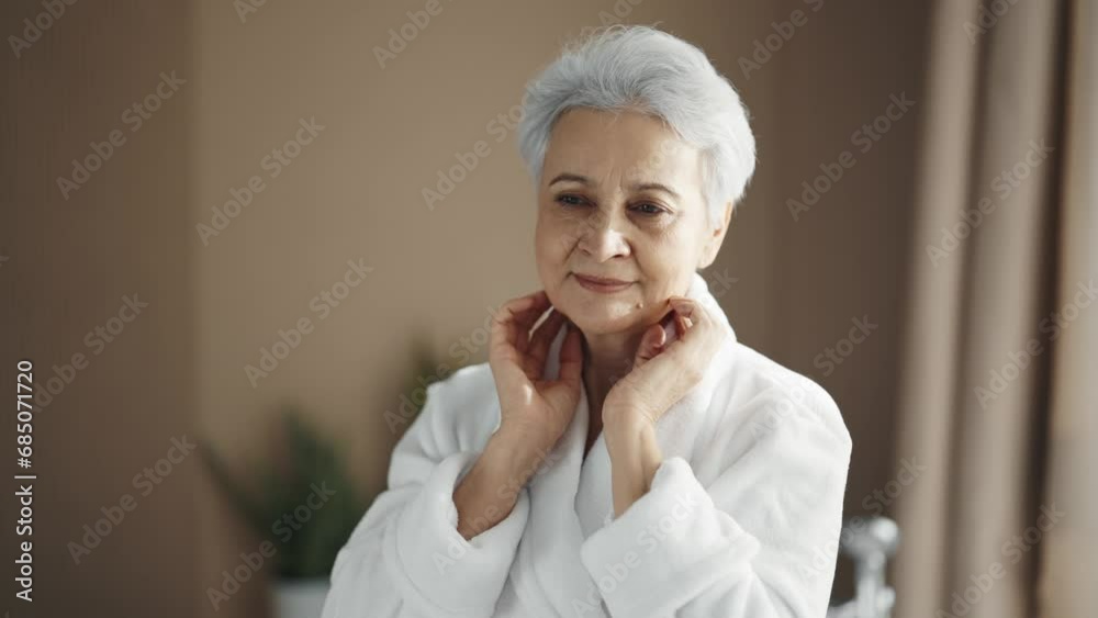 Positive senior woman in bathrobe straightens hair hairstyle rubbing massaging face looking at mirror at home. Morning routine, face skincare self-care, natural beauty aging, getting old concept.