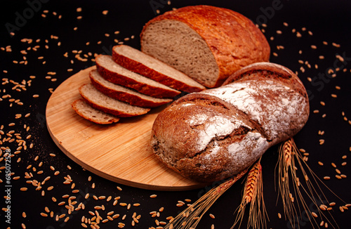 Whole and sliced breads, wheat ears and scattered wheat grains on wooden plate on black background. Photograph. (ID: 685072528)