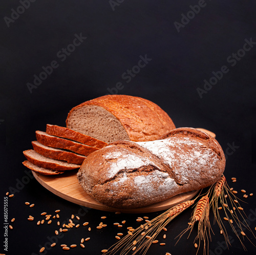 Whole and sliced breads, wheat ears and scattered wheat grains on wooden plate on black background. Photograph. (ID: 685075555)