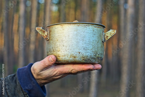 Fototapeta Naklejka Na Ścianę i Meble -  aluminum small dirty for cooking classic old with handles closed with a lid retro pan in black soot in the hand of a man during the day on the street