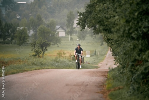 A young and fit cyclist on a bicycle explores the country landscape, riding on a road in the forest with a helmet and a mountain bike. Gravel biker on a path, enjoying the sport and the nature. © AlexGo