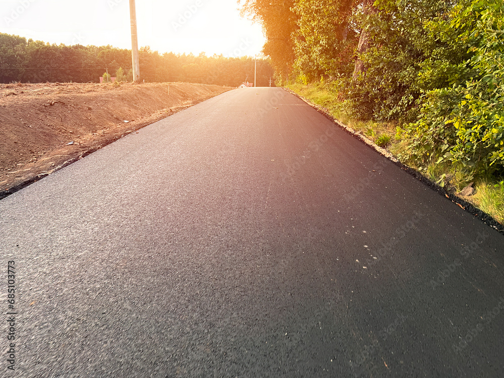 A close-up view of the hot and fresh asphalt layer on a newly constructed road, showing the texture and grain of the material and the equipment used for paving. construction and laying of the pavement - obrazy, fototapety, plakaty 