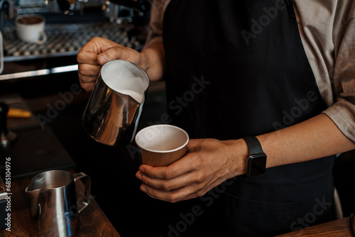 Close-up of a barista's hands pouring frothed milk into a paper coffee cup, with a blurred espresso machine in the background