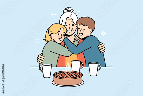 Happy family of grandmother and two little grandchildren hugging sitting at table with hot cake and mugs of tea. Smiling grandmother rejoices at meeting great-grandchildren after long absence