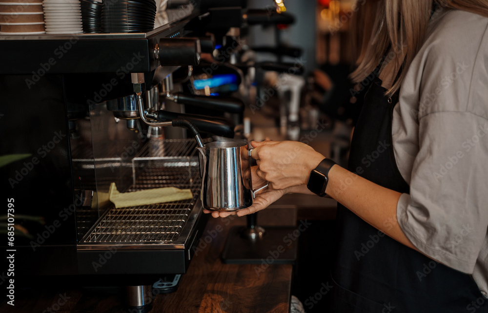 Close-up of a barista's hands pouring frothed milk into a paper coffee cup, with a blurred espresso machine in the background
