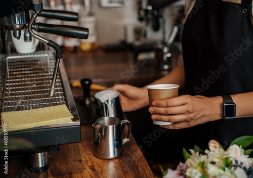 Close-up of a barista s hands pouring frothed milk into a paper coffee cup  with a blurred espresso machine in the background