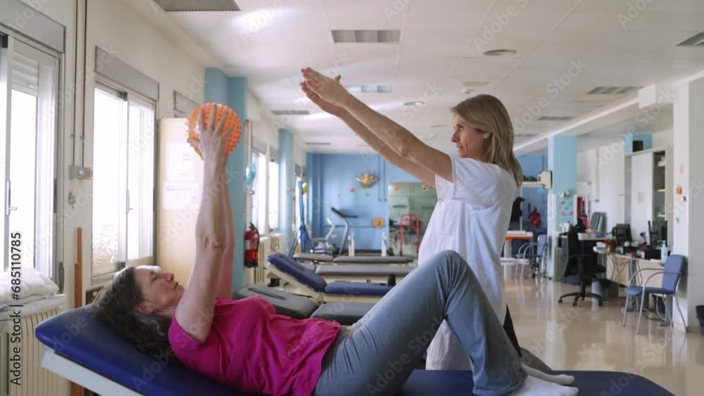 Blonde physiotherapist is showing to her patient how to do some exercises with a ball for recover her injury on her shoulders