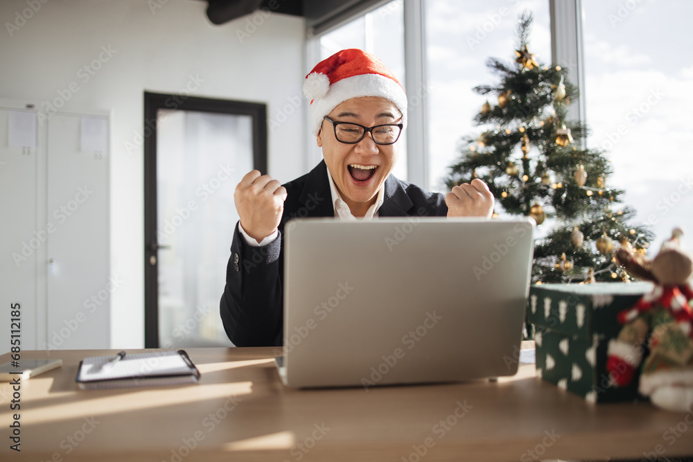 Emotional adult in Santa hat lifting fists of hands in excitement while looking at computer screen in office decorated with Christmas tree. Delighted asian male making successful holliday purchase Emotional adult in Santa hat lifting fists of hands in excitement while looking at computer screen in office decorated with Christmas tree. Delighted asian male making successful holliday purchase