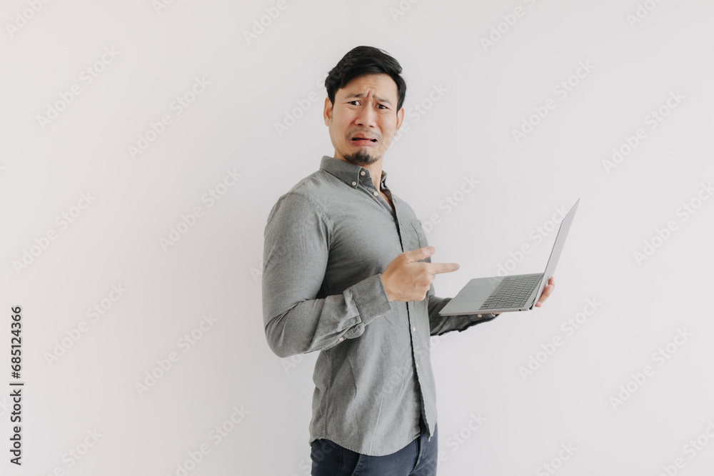 Asian man with beard wear grey shirt, disappointed and unsatisfied face, holding notebook hand pointing at laptop, unhappy funny face looking at camera isolated over white background wall.