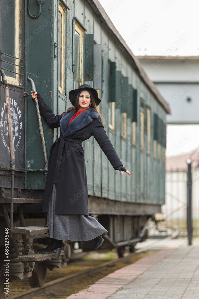 A beautiful girl in a black coat and hat near an old steam locomotive and big iron wheels. A young woman with long dark hair. Vintage portrait of the last century, retro journey.
