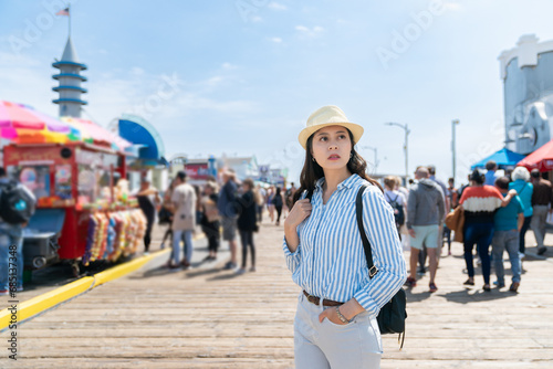 stylish asian Japanese girl backpacker with hand in pocket exploring busy santa monica pier near food carts under blue sky