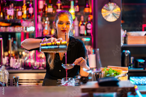 Beautiful barmaid with blonde hair looking at camera while preparing cocktail at pub photo