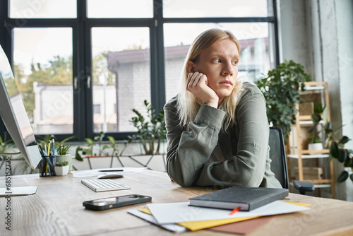 good looking non binary person in casual attire sitting at table with hand under chin, business photo