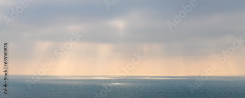 Pastel orange sunrays through grey clouds over the sea © Jeff