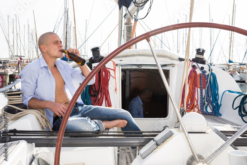 European guy with a naked torso and a bottle of beer in his hand sits on the deck of a yacht in the port