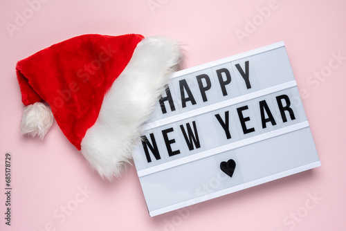Lightbox with text HAPPY NEW YEAR and Santa Claus hat on pastel pink background. Minimal Christmas or New Year concept.