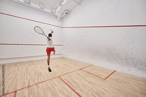 young and athletic african american player with racquet jumping and playing squash in court