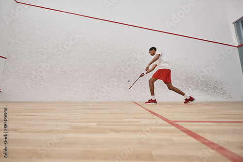 african american young man in red shorts holding racquet while playing squash inside of court