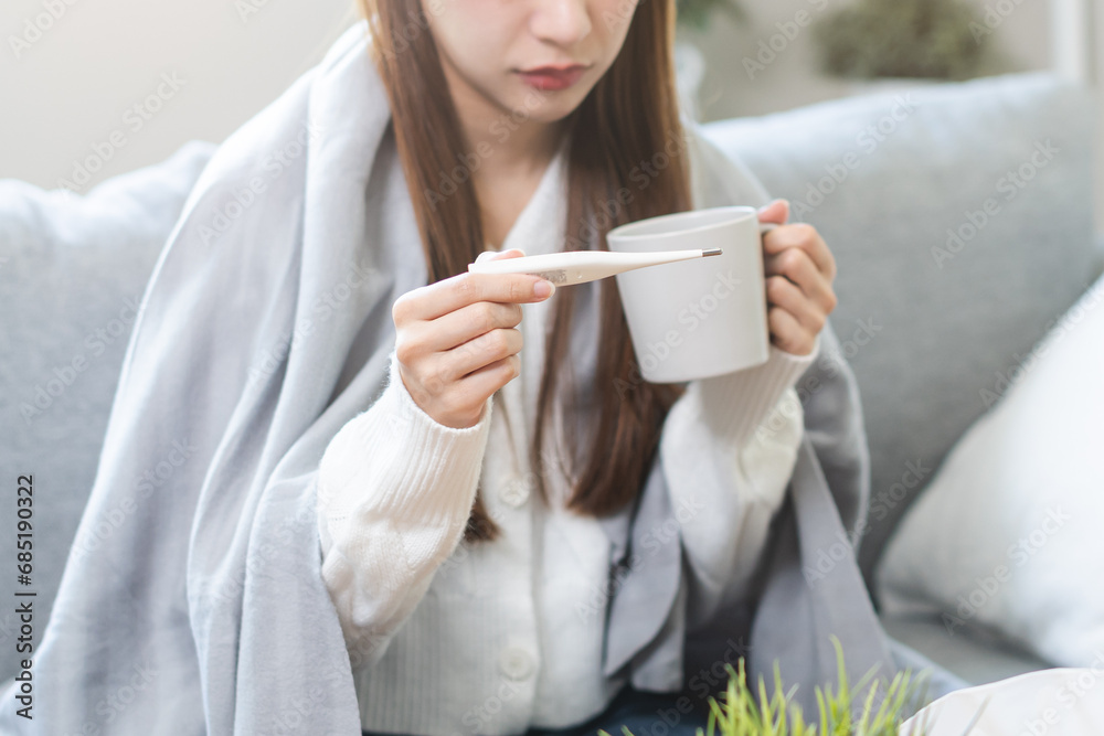 Sick, influenza asian young woman, girl have a fever, hand holding thermometer for check measure body temperature and drink hot water, illness while sitting rest on sofa at home. Health care concept. Sick, influenza asian young woman, girl have a fever, hand holding thermometer for check measure body temperature and drink hot water, illness while sitting rest on sofa at home. Health care concept.