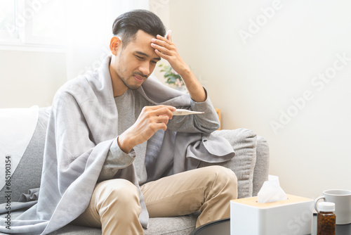 Sick, influenza asian young man, male have fever hand holding thermometer, touching forehead for check measure body temperature, illness while sitting rest on sofa at home. Health care on virus person