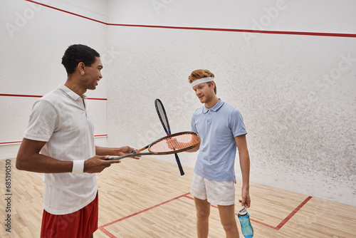 joyful interracial male friends with squash racquets and bottles with water chatting  in court © LIGHTFIELD STUDIOS