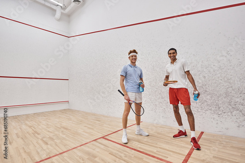 cheerful interracial sportsmen with squash racquets and bottles with water standing inside of court