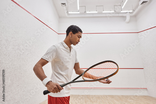 african american man in active wear holding squash ball and racquet while playing inside of court © LIGHTFIELD STUDIOS
