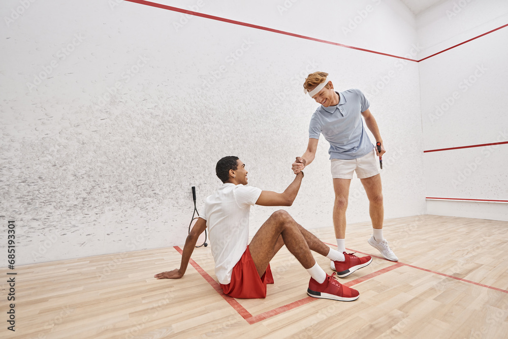 happy redhead man helping his african american friend to stand up from floor inside of squash court