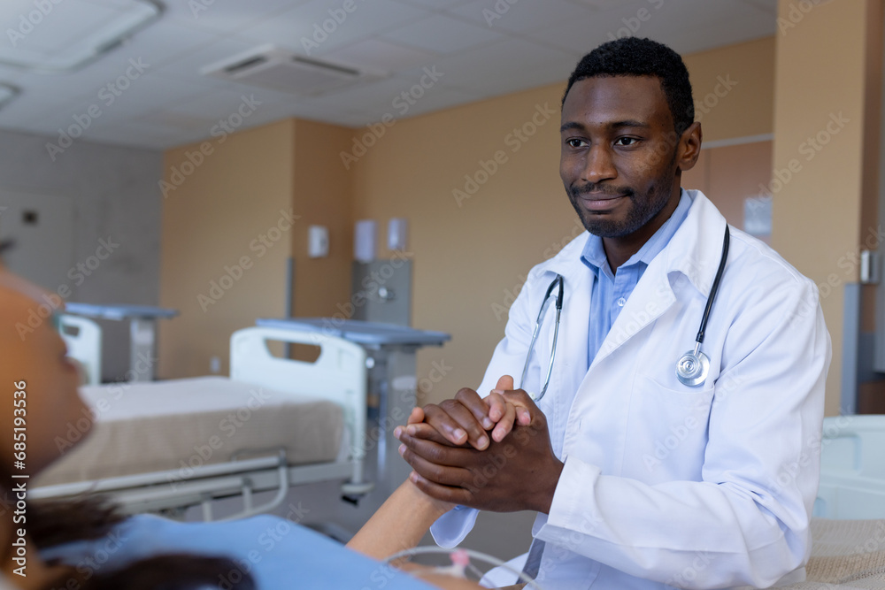 Diverse male doctor holding hand of female patient with iv drip in bed in hospital room