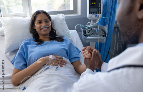 Happy diverse male doctor holding hand of female patient with iv drip in bed in hospital room photo