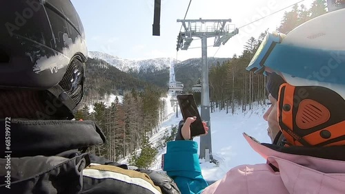 Man and a woman in winter sportswear, helmets and goggles ride on an open elevator at a ski resort, and take a selfie together on the phone against the backdrop of snow and forest. Winter holidays photo