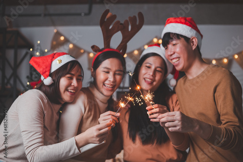 Group of young Asian man and women as friends having fun at a New Year's celebration, holding sparklers at a midnight countdown Party at home with Christmas tree decoration for holiday season.